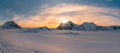 The sun sets behind the rugged, snow-covered peaks of Katla Glacier, casting a golden glow over the