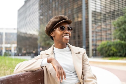 A fashionable black woman in sunglasses and a hat sits on a city bench, exuding confidence and