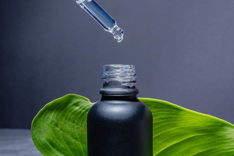 A dropper releasing a clear serum into a matte black bottle, set against a solid grey background