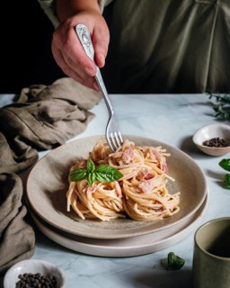 A hand elegantly twirls a fork in a creamy pasta dish topped with fresh basil leaves. The scene