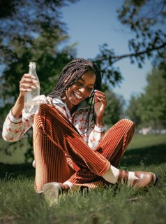 A joyful African American woman with braided hair laughs heartily while sitting on grass and