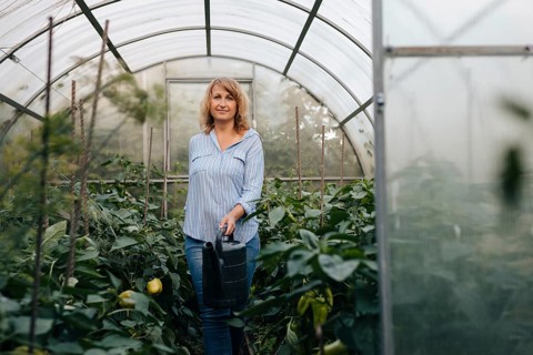 A woman stands in a lush greenhouse, holding a watering can. Surrounded by thriving plants, she