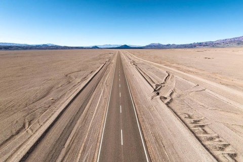 A straight road stretches across a vast desert landscape of the argentine Puna, leading into