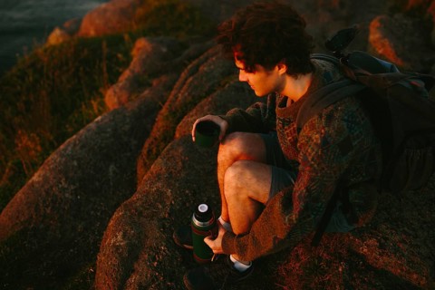 A young male hiker sits on rugged rocks at Cape Spear Lighthouse, watching the sunset. He is