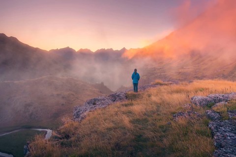 A lone person stands on a grassy ridge, gazing at misty mountains during dusk in summer. Warm light