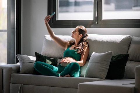 Pregnant woman sitting comfortably on a couch, wearing a yoga outfit, and taking a selfie. She