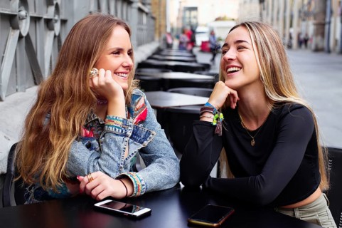 Two teenage girls smile and chat outdoors, with cell phones on the table, enjoying each other's