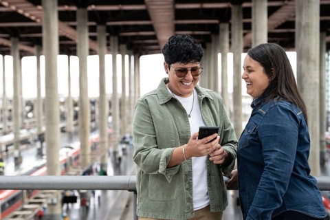 A joyful moment between a lesbian curvy couple at a train station, sharing smiles and a phone,