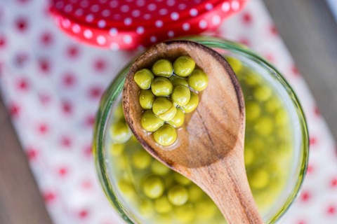 A wooden spoon holds green homemade organic peas over a glass jar, set on a white cloth with red