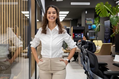 A woman in a white blouse stands confidently in a modern office. Her smile reflects a positive and