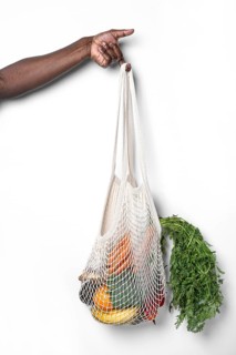 A hand holds a reusable cotton mesh bag filled with fresh vegetables and fruits, showcasing