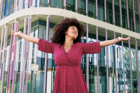 Young Arab woman with afro hair, wearing a pink dress, joyfully extends her arms in front of a