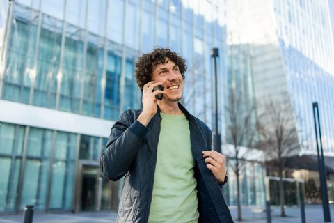 A businessman stands outside a sleek, modern glass building, smiling as he talks on his smartphone,