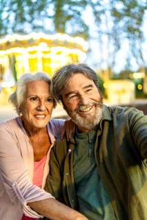 Senior couple taking selfie sitting on bench in the city at night next to carousel