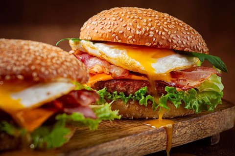 Two juicy burgers sit on a wooden board, each filled with lettuce, bacon, cheese, and fried eggs