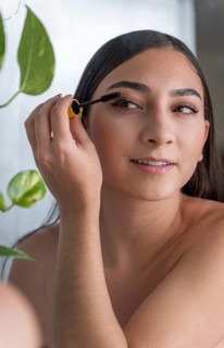 A woman smiling as she applies mascara in a brightly lit room. The presence of a vibrant green