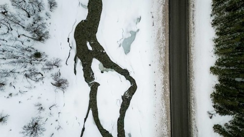 Aerial view of a Nordic landscape showcasing a road flanked by a frozen stream and snow-laden trees