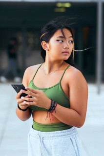 A teenage girl holds a smartphone while standing outdoors. Dressed in a green top and light pants,