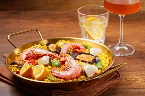 A large dish of seafood paella features shrimp, mussels, and other ingredients. A glass with a