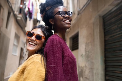 Two friends smiling joyfully, standing back-to-back in a narrow street. One wears sunglasses and a
