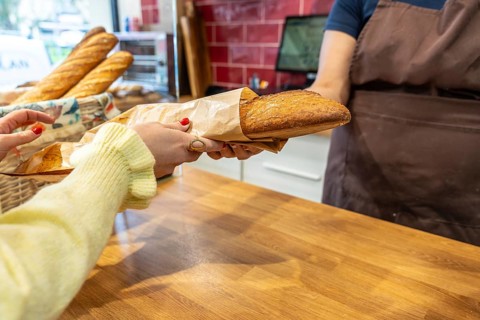 Customer hands collecting a freshly baked baguette wrapped in paper from a baker behind a wooden
