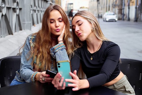 Two teenage girls sit together, using their cell phones and sharing a fun moment. They are