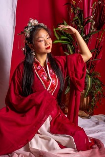 A woman in a red robe sits on a bed with a potted plant behind her. She is wearing a flower