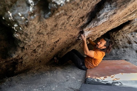 A climber in an orange shirt tackles a challenging boulder face in the wilderness, gripping rocks