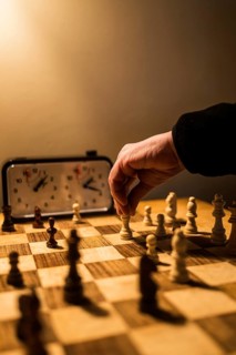 A hand moves a chess piece on a wooden board, lit warmly. An analog clock keeps time, highlighting