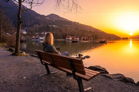 A person sitting on a bench by the lake at sunset with mountains in the background, Großer Alpsee,