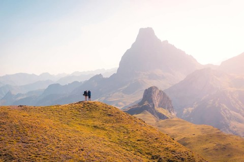 Two hikers stand on a grassy hill, gazing at the breathtaking mountain range in the Pyrenees. The
