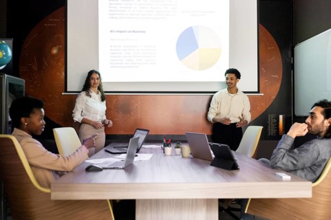 A diverse team engages in a modern office meeting. Two presenters stand by a screen displaying a