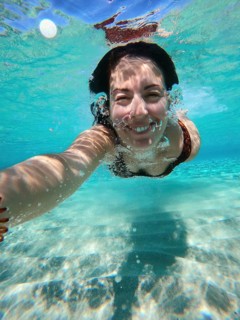 A woman swims underwater in Greece, capturing a selfie. The clear blue water reflects sunlight,