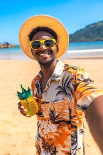 Young man taking a selfie on a tropical beach, wearing sunglasses and a straw hat, holding a
