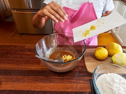A woman wearing a pink apron prepares lemon cookies in the kitchen. She adds lemon zest to a brown