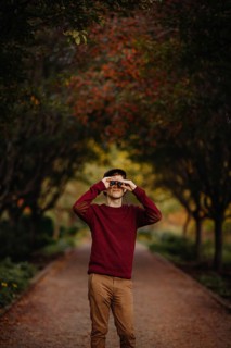 A man in a red sweater looks through binoculars amidst vibrant autumn foliage in a scenic park