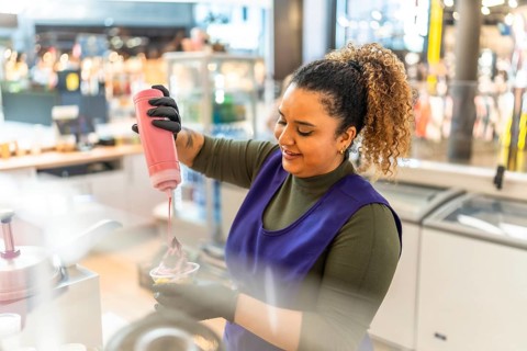 Woman employee in uniform smiling as she pours pink syrup over frozen yogurt in a cup at a shop