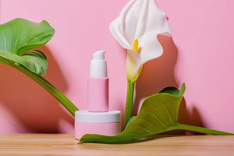 Pink skincare cosmetic products displayed with lush green leaves and a white calla lily against a