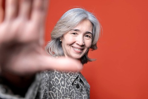 Asian senior woman smiles warmly while posing in front of a vibrant orange background. Her gray