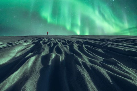 Unrecognizable person in red walks alone under the Northern lights on a snowy landscape at night,