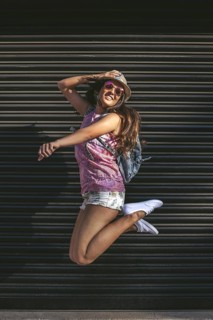 A young woman in sunglasses and a hat jumps joyfully against a dark urban background, embodying