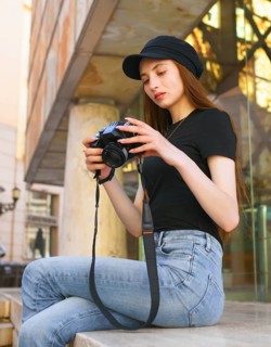 A woman photographer sits on a city bench, focused on her camera. Dressed in casual attire with a
