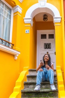 Vertical portrait of a smiling african woman using phone sitting in outside stairs of a yellow