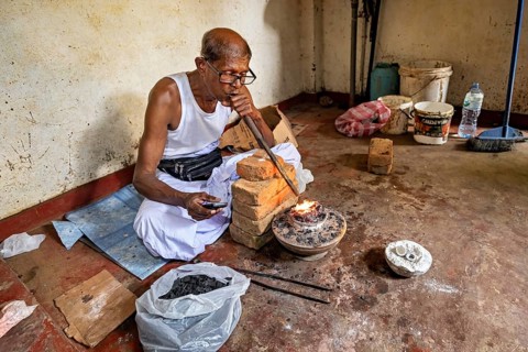 A man sits in a workshop, uses a bellows to start a fire, a man burns sapphires in a small oven in