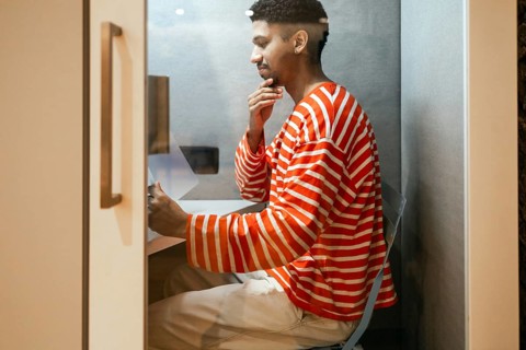 A man wearing a striped sweater works intently in a compact coworking phone booth, representing