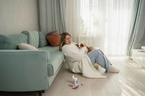 A woman cuddles her dog in a cozy home setting, sitting next to a teal sofa. Sunlight filters