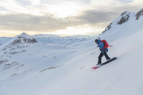 A snowboarder rides down a pristine snowy slope, surrounded by stunning alpine peaks under a soft,