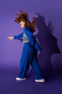 A young woman dances energetically against a vibrant purple background. Her dynamic pose and