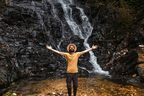 A young man stands with outstretched arms in front of a waterfall in the Choco Andino rainforest in