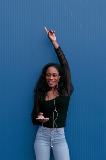 A young woman in casual attire, listening to music with earphones, raises her hand in excitement.